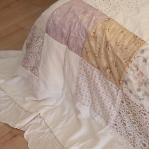 May include: A patchwork quilt featuring a variety of fabric squares in shades of white, cream, soft lavender, and muted tan. The quilt is adorned with floral and dotted patterns and is finished with a wide, gathered white fabric ruffle edge.