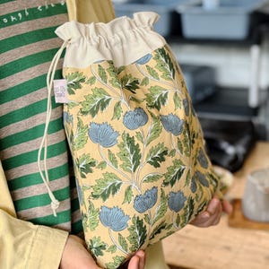 May include: A drawstring pouch featuring a yellow background with a repeating pattern of blue flowers and green leaves. The top section is a solid cream colour with a drawstring closure. A small pink tag with white text is attached to the side.