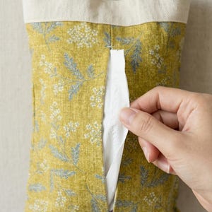Peut inclure: Un étui à mouchoirs en tissu jaune à motifs floraux, avec un dessus crème. Le couvercle comporte une fente pour la distribution des mouchoirs. Le tissu présente un motif de petites fleurs blanches et de feuilles grises.