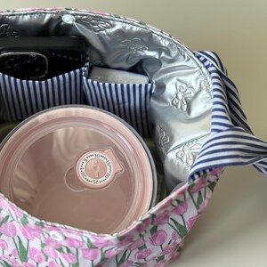 May include: A floral print lunch bag with a pink and white tulip pattern. The bag has a silver insulated lining and blue and white striped pockets. Inside, a pink container and a black phone are visible.