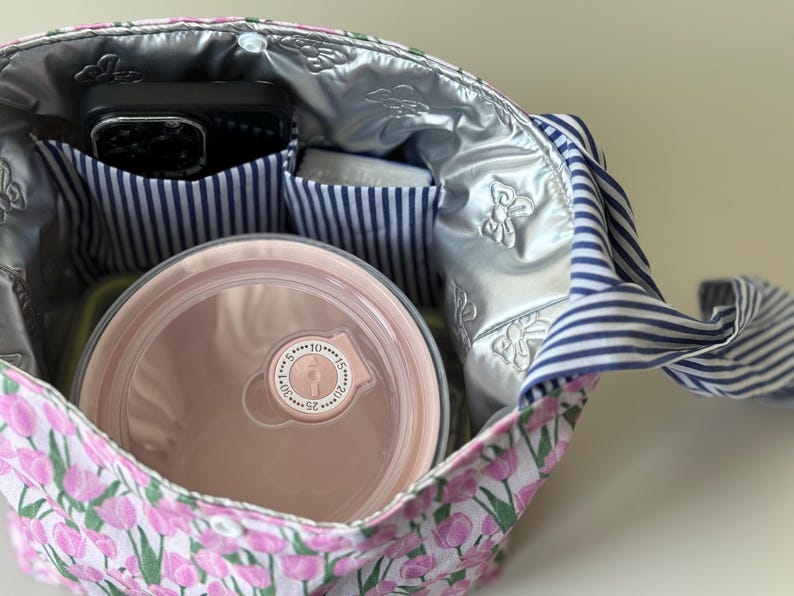 May include: A floral print lunch bag with a pink and white tulip pattern. The bag has a silver insulated lining and blue and white striped pockets. Inside, a pink container and a black phone are visible.