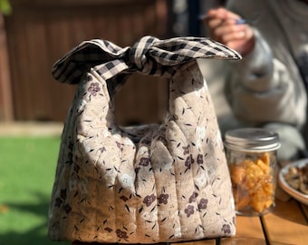 May include: A quilted lunch bag with a floral pattern in shades of beige and brown. The bag has a handle tied in a black and white checkered bow. A glass jar with snacks sits next to the bag on a wooden table.