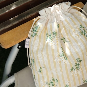 May include: A drawstring bag with a cream-colored top and a body featuring vertical yellow stripes and floral patterns. The bag is made of fabric and has a drawstring closure. The bag is resting on a wooden chair.