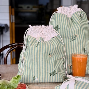 May include: Two green and white striped drawstring bags with embroidered floral details. The bags have white ruffled tops and white drawstrings. A glass of orange juice and a wrapped item are on the wooden table.