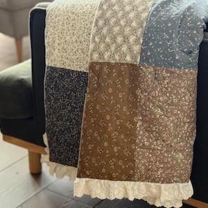 Puede incluir: Una colcha de retazos con paneles de tela cuadrados en varios patrones florales y colores apagados, incluyendo crema, azul marino, marrón y azul polvoriento. La colcha está terminada con un borde de encaje blanco con ojales.