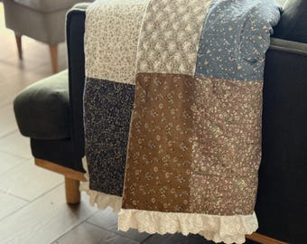 Op de afbeelding: Een patchwork quilt met vierkante stoffen panelen in diverse bloemmotieven en gedempte kleuren, waaronder crème, marineblauw, bruin en stofblauw. De quilt is afgewerkt met een witte kanten rand met oogjes langs de onderkant.