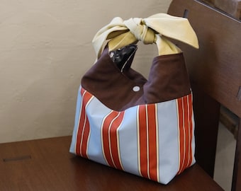 May include: A fabric handbag featuring a light blue and orange vertical striped pattern on the lower section, a solid brown upper section, and a large cream-colored fabric bow tied at the top handle. A small white snap button is visible on the front.