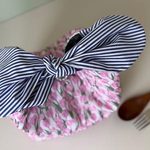 May include: A floral print bag with a large navy and white striped bow. The bag features a pink and white tulip pattern. The bow is tied in a knot, adding a decorative element to the bag's design.