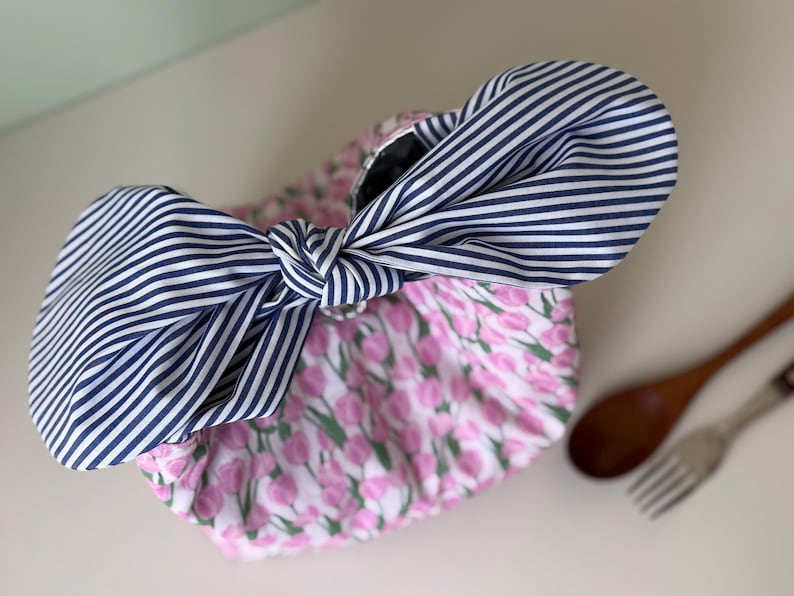 May include: A floral print bag with a large navy and white striped bow. The bag features a pink and white tulip pattern. The bow is tied in a knot, adding a decorative element to the bag's design.
