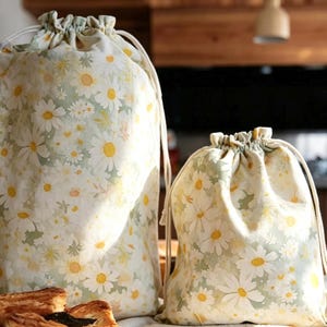 May include: Two drawstring bags with a daisy floral pattern. The bags are white with yellow and green daisy flowers. The bags have a drawstring closure and are made of fabric. One bag is larger than the other.