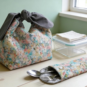 May include: A floral print lunch bag with a grey handle and a matching utensil case. A clear container with a white cloth and a spoon and fork are also visible. The bag and case have a colourful floral pattern.
