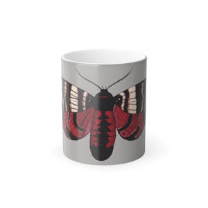Puede incluir: Taza de cerámica con un diseño de polilla. La polilla tiene alas a rayas blancas y negras con detalles rojos y un cuerpo negro, sobre un fondo gris. El interior de la taza es blanco.
