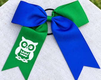 May include: A large hair bow featuring a split design of royal blue and bright green grosgrain ribbon. The bow is attached to a black elastic hair tie. A white graphic of an owl is printed on the green ribbon tail of the bow.