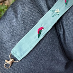 May include: A pale blue strap featuring embroidered parrots in red and green, and white flowers. The strap has a gold-coloured clasp and ring. The strap rests on grey fabric.