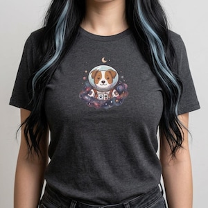May include: Dark gray t-shirt featuring a cartoon dog in an astronaut helmet, surrounded by a colorful nebula and a crescent moon. The dog has brown and white markings. The design is centered on the front of the shirt.