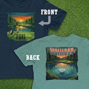 May include: Two golf-themed t-shirts. The front shirt is navy blue with a golf course scene and the word "PURE". The back shirt is teal with a golf course scene and the word "MULLIGAN". The shirts are on a green surface.