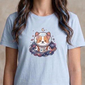 May include: Light blue t-shirt featuring a cartoon corgi in an astronaut helmet, floating in a colorful nebula. The design includes stars, a crescent moon, and a space-themed graphic. The shirt is made of a soft material.