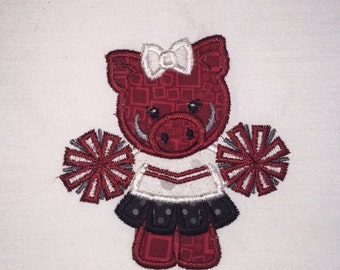 Peut inclure: Patch brodé représentant un cochon rouge portant une tenue de pom-pom girl blanche avec une jupe noire et blanche et tenant deux pompons.