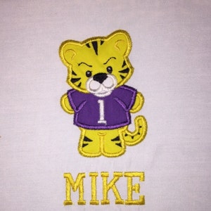 May include: Embroidered design of a yellow tiger wearing a purple jersey with the number 1 on it. The name Mike is embroidered below the tiger in yellow.