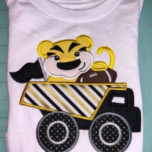 May include: White t-shirt with an embroidered design of a cartoon tiger driving a yellow and black dump truck. The tiger is wearing a brown football helmet and holding a football. The name "Andrew" is embroidered below the truck.
