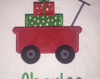 May include: A red wagon with a handle and wheels, carrying a stack of wrapped gifts. The top gift is red with white polka dots, the middle gift is green with white polka dots, and the bottom gift is green with white polka dots. The name "Charles" is written in green below the wagon.