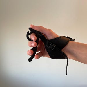 May include: Black archery release aid with a wrist strap. The release aid features a curved trigger and a leather wrist strap with a buckle for secure attachment. The device is designed for use with a bow and arrow.