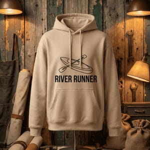 Puede incluir: Una sudadera con capucha beige con un bolsillo delantero y un cordón. La sudadera presenta un gráfico negro de un kayak y remos, con el texto "RIVER RUNNER" debajo. El fondo es una pared de madera.