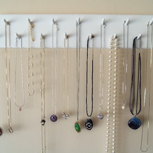 May include: A white wooden necklace organizer with multiple hooks, displaying various necklaces. The necklaces vary in style, including delicate chains, pearl strands, and pendants with gemstones and charms. The background is a neutral wall.