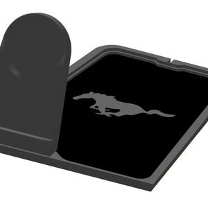 Puede incluir: Soporte negro para teléfono de coche con un emblema gris de un caballo corriendo sobre una superficie negra. El soporte tiene un soporte vertical y una base rectangular.