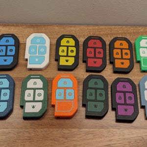 Puede incluir: Doce fundas rectangulares para llaves dispuestas en dos filas. Cada funda presenta una disposici&oacute;n de cinco botones con iconos de bloqueo, desbloqueo, encendido y alarma. Las fundas vienen en varios colores s&oacute;lidos, incluyendo azul, blanco, amarillo, rojo, naranja, verde, azul oscuro, turquesa y morado.