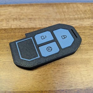 Puede incluir: Una funda negra texturizada para llave con tres botones de color azul claro. El bot&oacute;n superior muestra un icono de candado abierto, el central una flecha circular y el bot&oacute;n derecho un icono de candado cerrado.
