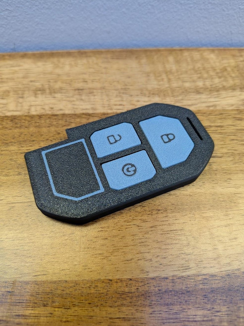 Puede incluir: Una funda negra texturizada para llave con tres botones de color azul claro. El bot&oacute;n superior muestra un icono de candado abierto, el central una flecha circular y el bot&oacute;n derecho un icono de candado cerrado.