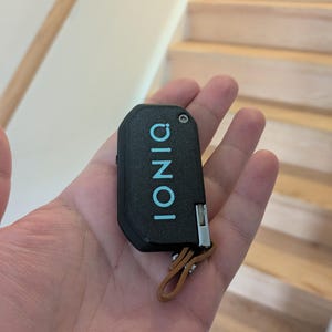Puede incluir: Un llavero de coche negro con textura que presenta la palabra IONIQ impresa en letras may&uacute;sculas de color azul claro. El dispositivo rectangular tiene una peque&ntilde;a bisagra de metal plateado en el lateral y una trabilla de cuero marr&oacute;n.