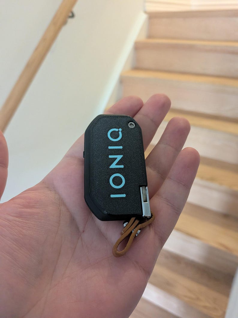 Puede incluir: Un llavero de coche negro con textura que presenta la palabra IONIQ impresa en letras may&uacute;sculas de color azul claro. El dispositivo rectangular tiene una peque&ntilde;a bisagra de metal plateado en el lateral y una trabilla de cuero marr&oacute;n.
