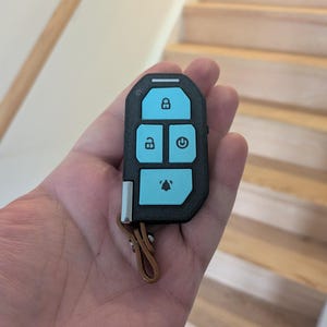 Puede incluir: Un mando electr&oacute;nico negro con acabado texturizado y cuatro botones de color azul claro. Los botones muestran iconos de bloqueo, desbloqueo, encendido y alarma. Una peque&ntilde;a trabilla de cuero marr&oacute;n est&aacute; sujeta a la parte inferior.