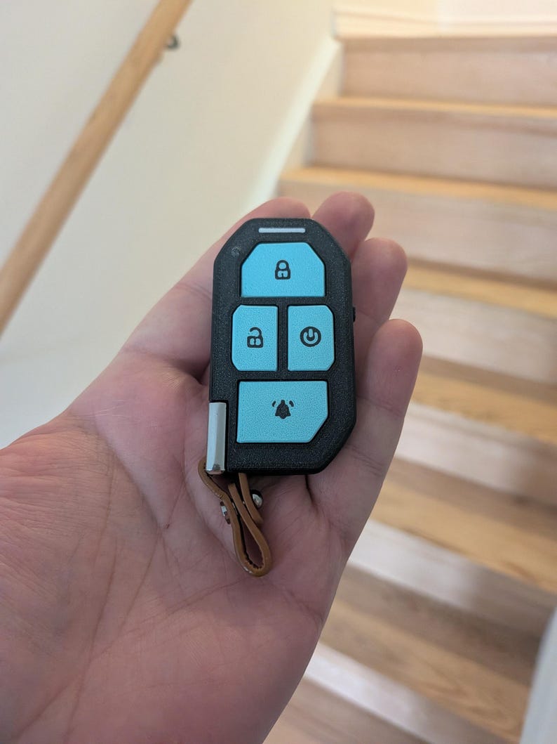 Puede incluir: Un mando electr&oacute;nico negro con acabado texturizado y cuatro botones de color azul claro. Los botones muestran iconos de bloqueo, desbloqueo, encendido y alarma. Una peque&ntilde;a trabilla de cuero marr&oacute;n est&aacute; sujeta a la parte inferior.