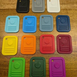 Puede incluir: Una colecci&oacute;n de catorce etiquetas rectangulares de pl&aacute;stico dispuestas en filas. Cada etiqueta presenta un centro circular elevado, tres l&iacute;neas horizontales en la parte inferior y un peque&ntilde;o orificio en la parte superior. Las etiquetas son de color negro, gris, blanco, azul claro, azul, azul oscuro, amarillo, naranja, rojo, granate, verde, verde oscuro y morado.