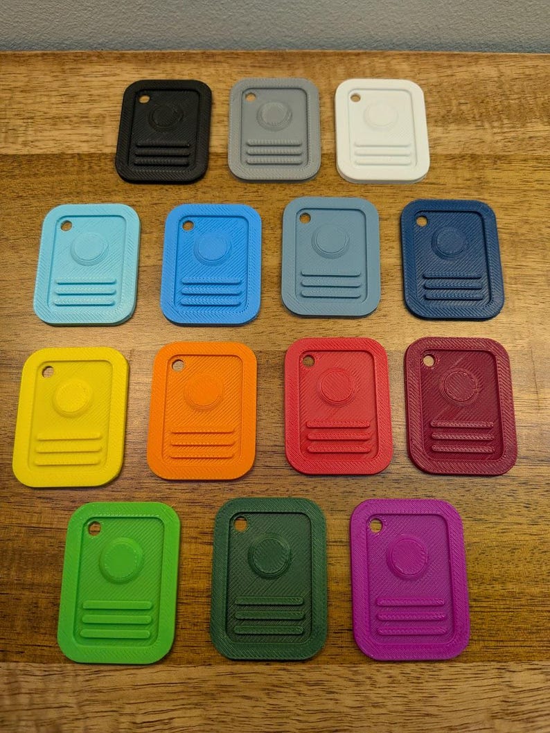 Puede incluir: Una colecci&oacute;n de catorce etiquetas rectangulares de pl&aacute;stico dispuestas en filas. Cada etiqueta presenta un centro circular elevado, tres l&iacute;neas horizontales en la parte inferior y un peque&ntilde;o orificio en la parte superior. Las etiquetas son de color negro, gris, blanco, azul claro, azul, azul oscuro, amarillo, naranja, rojo, granate, verde, verde oscuro y morado.