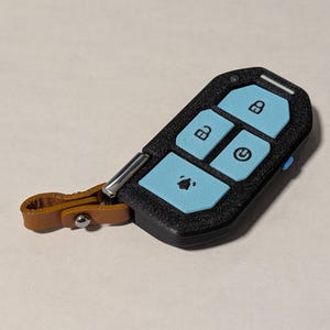 Puede incluir: Una funda negra texturizada para llave con botones azul claro que muestran iconos de bloqueo, desbloqueo, encendido y alarma. El dispositivo incluye un peque&ntilde;o punto de sujeci&oacute;n met&aacute;lico plateado unido a una trabilla de cuero beige con broche.