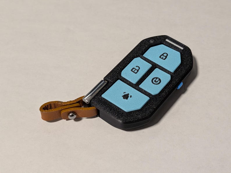 Puede incluir: Una funda negra texturizada para llave con botones azul claro que muestran iconos de bloqueo, desbloqueo, encendido y alarma. El dispositivo incluye un peque&ntilde;o punto de sujeci&oacute;n met&aacute;lico plateado unido a una trabilla de cuero beige con broche.