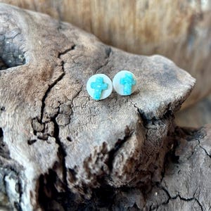 Peut inclure: Boucles d'oreilles à clous avec un motif de croix turquoise sur une base circulaire blanche. Les boucles d'oreilles sont présentées sur un morceau de bois vieilli, mettant en valeur leur couleur et leur texture.