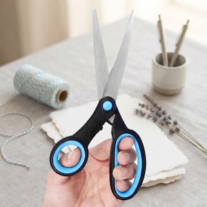 May include: A pair of scissors with sharp, silver-colored metal blades and black handles featuring bright light blue rubberized inner grips. The central pivot point is secured with a matching light blue circular cap, providing a functional design.