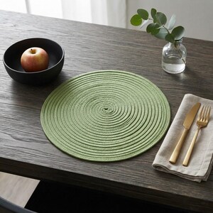 May include: A round, sage green woven rope placemat with a spiral pattern. Nearby is a black bowl containing a red apple, a clear glass vase with a green eucalyptus sprig, and a beige linen napkin with a gold-toned knife and fork set.