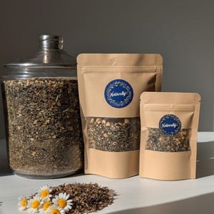 May include: An assortment of herbal tea products. A large glass jar and two kraft paper pouches with clear windows display the tea blend. The pouches have blue circular labels with the text "Naturally!". A small pile of tea and chamomile flowers are in the foreground.