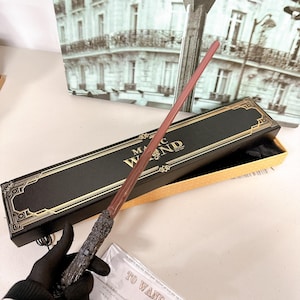 May include: A magic wand with a dark brown handle and a reddish-brown shaft, presented in a black box with gold lettering that reads "MAGIC WAND". The box is open, revealing a yellow interior. The wand is approximately 13 inches long.