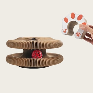 May include: A cat toy set featuring a corrugated cardboard play structure with a red ball inside and a separate arched scratcher. The scratcher has orange paw prints and a cat silhouette.