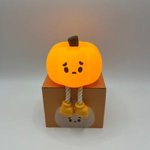 May include: An orange pumpkin-shaped decorative light with a sad face, brown stem, and yellow feet. The light sits on a brown box with a pumpkin face design. The legs are made of rope.