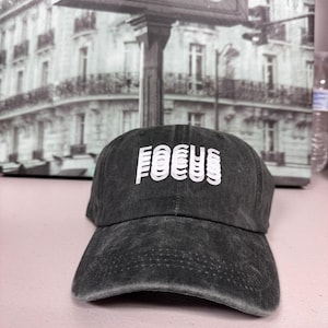 Peut inclure: Casquette de baseball gris foncé avec le mot "FOCUS" en blanc superposé sur le devant. La casquette a une visière incurvée et un aspect légèrement délavé. En arrière-plan, une image en noir et blanc d'un panneau "METRO".