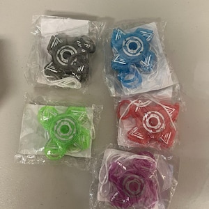 May include: Five fidget spinners in individual plastic packaging. The spinners are in various colors: black, blue, green, red, and purple. Each spinner has a central circular design with radiating arms and a white string attached.