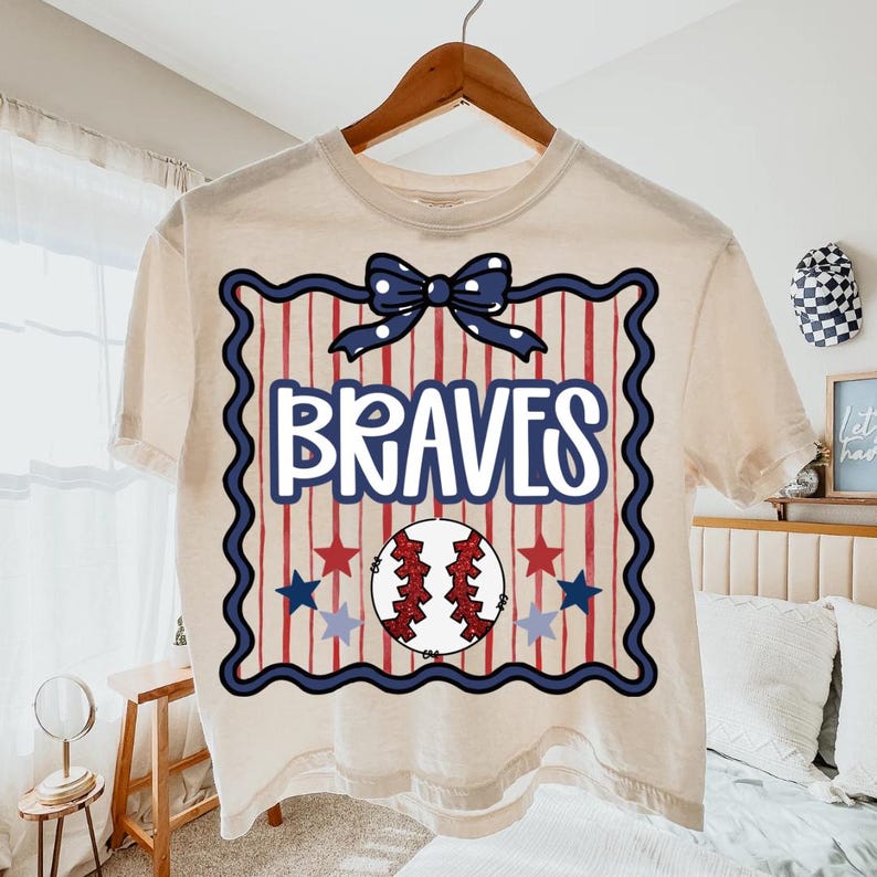 Puede incluir: Una camiseta de color crema con un gr&aacute;fico tem&aacute;tico de b&eacute;isbol. El dise&ntilde;o presenta la palabra "BRAVES" en blanco, una pelota de b&eacute;isbol, estrellas rojas y azules y un lazo azul. La camiseta est&aacute; colgada en una percha de madera.